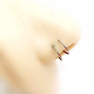 Può includere: Primo piano di un naso con due anelli al naso color argento. Gli anelli sono circolari e sembrano essere in metallo. L'incarnato è chiaro e lo sfondo è bianco.