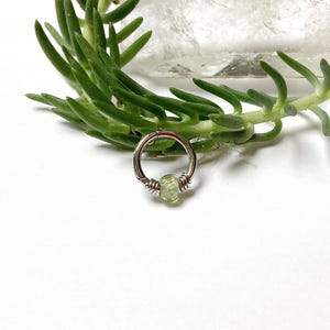 Puede incluir: Un aro de nariz plateado con una pequeña cuenta de piedra preciosa verde claro en el centro. El aro es circular y tiene un diseño simple y elegante. La piedra preciosa está envuelta con alambre de plata. El fondo incluye hojas suculentas verdes y un cristal claro.