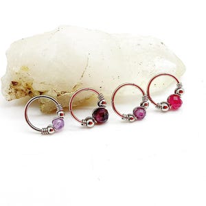 Peut inclure: Quatre boucles d'oreilles créoles argentées avec des pierres précieuses colorées. Chaque boucle d'oreille a un petit cercle avec une pierre précieuse et des perles argentées. Les pierres sont violettes, rouge foncé et roses.