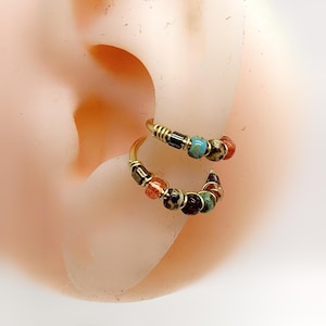 Peut inclure: Deux boucles d'oreilles créoles dorées, chacune ornée d'une série de perles colorées. Les perles comprennent des nuances de turquoise, de rouge, de noir et de marron. Les boucles d'oreilles sont présentées sur une oreille.