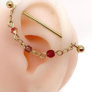 Könnte beinhalten: Goldfarbener Industrial-Barbell-Ohrring mit einer Kette aus kleinen, roten Perlen. Die Kette ist an einem Ende mit einer kleinen goldenen Kugel und am anderen Ende mit einer größeren goldenen Kugel verbunden. Der Ohrring wird an einem Ohr gezeigt.