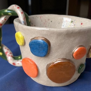 Puede incluir: Taza de cerámica hecha a mano con un acabado beige moteado y lunares de colores en relieve. El asa tiene un diseño retorcido con secciones verdes y blancas, salpicadas de rojo. Una taza de café o té única y artística.