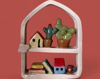 May include: A white ceramic shelf shaped like a house, featuring miniature ceramic items. The shelf holds a yellow house, cacti in pots, books, a blue house, and a small mug. The background is a solid red.