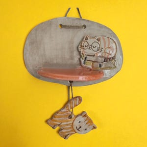 May include: A ceramic wall shelf featuring a sleeping cat design. The oval-shaped shelf has a small orange shelf and a hanging cat-shaped ornament. The cat on the shelf wears glasses and rests on books. The color scheme includes grey, orange, and cream.