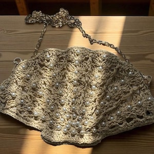 May include: A beige, seashell-shaped handbag with a chain strap. The bag is made of woven material and adorned with numerous white, pearl-like beads. The bag is lying on a wooden surface, with sunlight casting shadows.