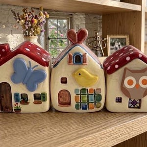 Può includere: Tre statuine in ceramica a forma di casa con tetti rossi e disegni stravaganti. Uno presenta una farfalla, un altro un uccello e l'ultimo un gufo. Sono esposte su una mensola di legno con altri oggetti decorativi.