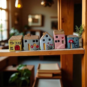 May include: A collection of miniature ceramic houses in various colors, including yellow, gray, blue, and pink, with decorative details like flowers, a heart, and a red door. One house has the word "tea" on it. The houses are arranged on a wooden shelf.