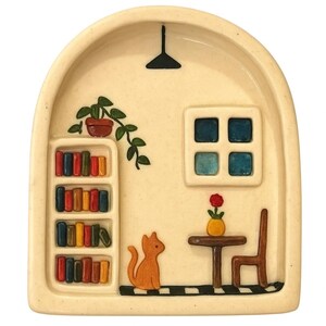 May include: A cream-colored ceramic tray with an arched top, featuring a miniature room scene. It includes a bookshelf with colorful books, a cat, a table, a chair, a window, and a hanging light. A small plant and a flower add detail.