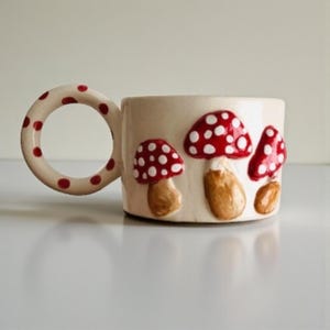 May include: Cream-colored ceramic mug with a large, round handle dotted with red spots. The mug features three-dimensional mushroom designs with red caps and white spots. The mushroom stems are brown.