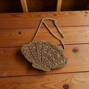 May include: A handmade, seashell-shaped handbag in a light brown color. The bag is crocheted with a detailed pattern and features a matching strap. The bag is resting on a wooden surface.
