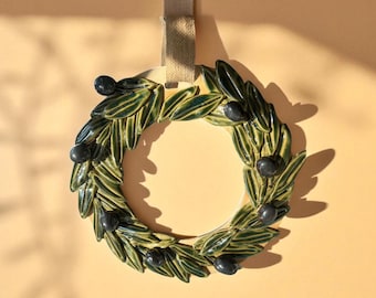 May include: Ceramic olive wreath with green leaves and black olives. The wreath is circular and has a beige ribbon for hanging. The background is a solid beige color.