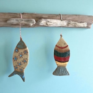 Puede incluir: Dos adornos de peces de cerámica colgados de un trozo de madera flotante. Un pez es de color beige con detalles florales azules, el otro es de color beige con rayas rojas, verdes y marrones. El fondo es azul claro.