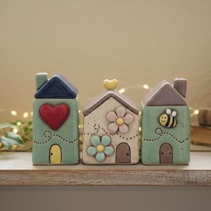 Könnte beinhalten: Drei dekorative Keramikhausfiguren mit detaillierten Designs. Eine hat ein rotes Herz, eine andere Blumen und die dritte eine Biene. Jedes hat einen Schornstein, eine Tür und ein kontrastierendes Dach. Die Häuser stehen auf einem Holzregal.