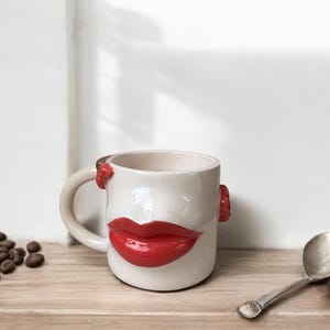 Pode incluir: Caneca de cerâmica branca com design de lábios vermelhos. A caneca tem uma alça curva e pequenos detalhes vermelhos nas laterais. A caneca está em uma superfície de madeira com grãos de café e uma colher.