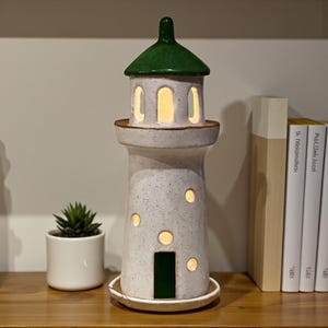 Puede incluir: Lámpara de cerámica con forma de faro, con tejado verde y cuerpo blanco moteado. La lámpara tiene varias aberturas redondas y arqueadas que emiten un brillo cálido. Una pequeña suculenta en una maceta blanca está cerca.