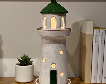 May include: A ceramic lighthouse-shaped lamp with a green roof and a white speckled body. The lamp has several round and arched openings that emit a warm glow. A small succulent in a white pot sits nearby.