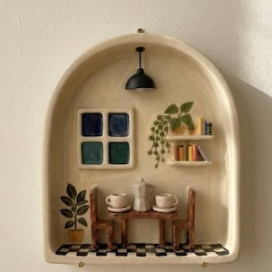 Puede incluir: Una obra de arte mural de cerámica que representa una escena de café en miniatura. Cuenta con una mesa con dos sillas, tazas de café y una cafetera. Hay una ventana, una lámpara colgante y una estantería con libros y plantas. El suelo tiene un patrón de cuadros blancos y negros.