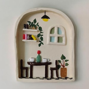 Puede incluir: Una pieza de arte mural de cerámica que representa una escena de café en miniatura. Presenta una mesa con dos tazas, sillas, una ventana, una lámpara colgante y una estantería con libros y plantas. La paleta de colores es principalmente crema, marrón y verde.