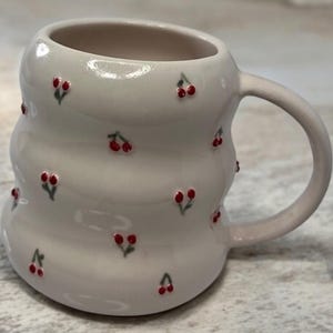 Puede incluir: Una taza de cerámica gris claro con un diseño ondulado único. La taza está adornada con pequeñas cerezas rojas pintadas a mano con tallos verdes. El asa es de color gris claro a juego. Una adición encantadora a cualquier cocina.