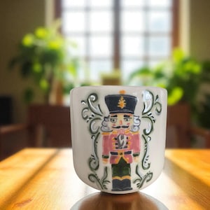 May include: A white ceramic mug featuring a detailed illustration of a nutcracker soldier in a pink coat, black hat, and green pants. The mug has decorative green swirls and is set on a wooden table. The background is blurred.