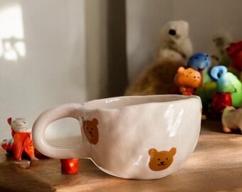 May include: A white ceramic mug with a bear design. The mug has a rounded shape and a large handle. Small bear illustrations are printed on the mug. The mug is on a wooden surface with small figurines in the background.