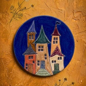 Può includere: Un piatto in ceramica con una scena di villaggio stravagante con edifici colorati e tetti a punta, su uno sfondo blu scuro. Il piatto è rotondo e ha una finitura lucida, con uno sfondo strutturato marrone dorato.