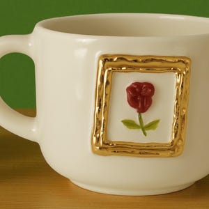 Puede incluir: Taza de cerámica color crema con un relieve enmarcado en oro de una rosa roja. La taza tiene forma redondeada y un asa curva. La rosa tiene hojas verdes y está centrada en la taza.