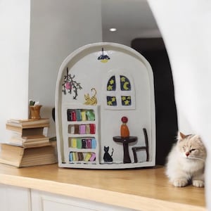 Puede incluir: Una caprichosa escultura de cerámica de una estantería con temática de gatos. La estructura blanca presenta una estantería con libros coloridos, una mesa, una silla y una ventana con diseños de luna y estrellas. También se representa un gato amarillo y un gato negro.