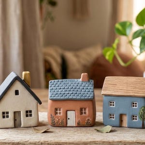 May include: Three miniature ceramic houses in cream, orange, and blue with grey, blue, and wood-tone roofs. Each house has small windows and doors. The houses are arranged on a wooden surface, with a plant in the background.