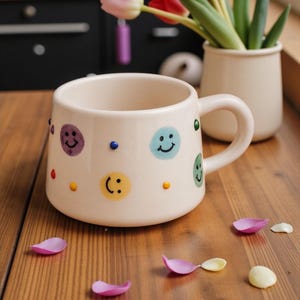 May include: A cream-colored ceramic mug adorned with colorful smiley faces and dots. The mug has a curved handle and sits on a wooden surface, with scattered flower petals and a vase of tulips in the background. A cheerful and whimsical design.