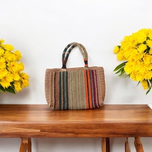 May include: A hand-crocheted tote bag with vertical stripes in shades of brown, green, orange, and burgundy. The bag has matching striped handles. The bag is sitting on a wooden surface, with yellow flowers on either side.