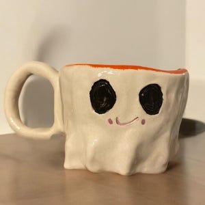Könnte beinhalten: Eine handgefertigte Keramiktasse mit einem geisterhaften Design. Die cremefarbene Tasse hat zwei große schwarze Augen, ein kleines Lächeln und einen orangefarbenen Rand. Die Tasse hat einen abgerundeten Henkel und steht auf einer Holzoberfläche.