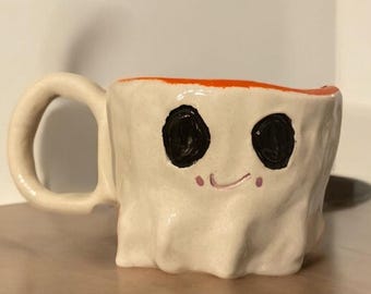 May include: A handmade ceramic mug with a ghost-like design. The off-white mug features two large black eyes, a small smile, and an orange rim. The mug has a rounded handle and sits on a wooden surface.