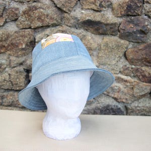 Peut inclure: Un chapeau de pêcheur en jean bleu clair avec un large bord. Le chapeau présente un patch carré avec un motif floral coloré. Le chapeau est présenté sur une tête de mannequin blanche contre un mur de pierre.