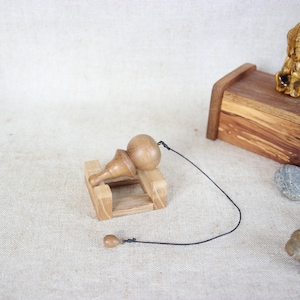 Op de afbeelding: Een houten slinger met een ronde bal aan een touwtje, rustend op een houten standaard. De slinger bevindt zich op een lichtbruin stoffen oppervlak met een kleine houten doos en een paar stenen.