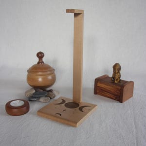 Op de afbeelding: Een houten standaard met een vierkante basis met maansikkels en cirkelontwerpen. Een kleine houten kist, een kaarsenhouder en een houten pot zijn ook aanwezig. De items zijn gerangschikt op een wit oppervlak.