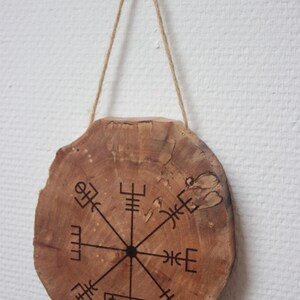 Peut inclure: Une tranche de bois ronde avec un motif de boussole noir, comprenant un point central et huit symboles directionnels. La tranche est suspendue à une ficelle de ficelle.