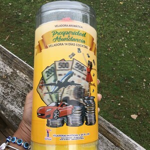 May include: A cylindrical yellow candle in a clear plastic container. The label reads "Prosperidad Abundancia" with images of money, a red car, and an airplane. The candle is made by Veladoras Misticas.