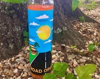 May include: A glass candle with an orange wax top and a blue and yellow label. The label features a road with a white line down the center, a house, a sun, and clouds. The text on the label reads "ROAD OPEN", "SUCCESS", "HEALTH", and "BUSINESS".