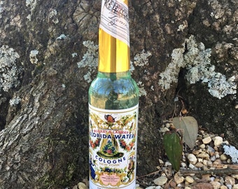 Może przedstawiać: Przezroczysta szklana butelka z etykietą z złotej folii. Etykieta przedstawia kwiatowy wzór i tekst "Murray & Lanman's Florida Water Cologne New York". Butelka stoi na podłożu z małych kamieni.