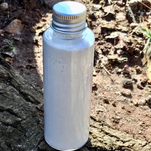 May include: A small white bottle with a silver lid, filled with a white powder. The bottle is sitting on a piece of tree bark.