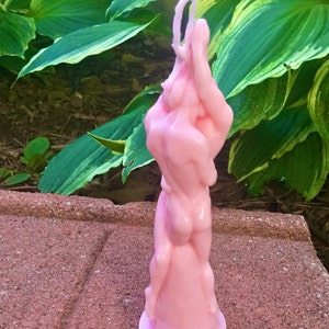 May include: A pink candle shaped like two figures embracing. The candle has a white wick and is sitting on a brown surface.