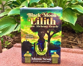 May include: A 44-card deck and guidebook for the Black Moon Lilith Cosmic Alchemy Oracle. The cover features a black and green illustration of a woman with a blue and green scarf. The text on the cover reads "Black Moon Lilith Cosmic Alchemy Oracle" and "Adama Sesay".