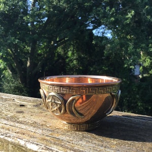 May include: A copper bowl with a decorative design featuring a pentacle, crescent moons, and a Greek key pattern. The bowl is sitting on a wooden surface.