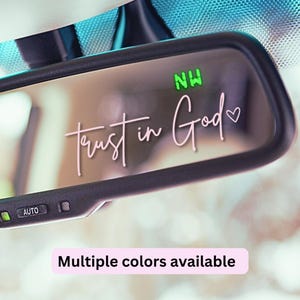 May include: A car's rearview mirror with the words "Trust in God" in a delicate pink script. The mirror also displays the letters "NW" in green. The bottom of the image reads "Multiple colors available".