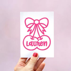 May include: A white rectangular card featuring a bright pink graphic of two cherries connected by a bow at the top. The name Lauren is written in a bold, rounded pink font across the center of the two cherry shapes in a clean design.