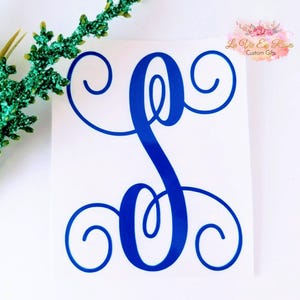 May include: A blue vinyl decal featuring a large, stylized letter S with decorative, swirling flourishes at the top and bottom. The decal is applied to a white surface, and a small logo with the text La Vie En Roses Custom Gifts is visible.
