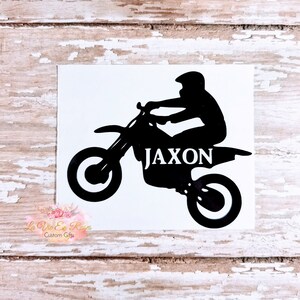 May include: A black vinyl decal featuring a silhouette of a person riding a dirt bike. The name JAXON is printed in white capital letters across the body of the motorcycle. The decal is presented on a white rectangular backing sheet.