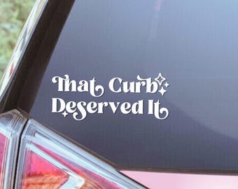 May include: A white vinyl decal applied to a car window featuring the text "That Curb Deserved It" in a stylized, retro-inspired font. The design includes small four-pointed star accents positioned near the lettering on a dark glass surface.