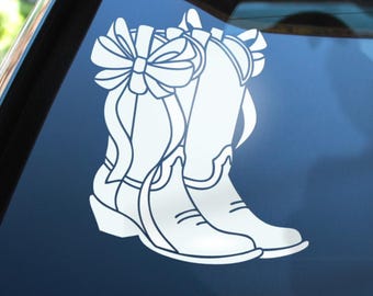 May include: A white vinyl decal featuring a pair of cowboy boots adorned with a large decorative bow on the side. The design is rendered in clean, solid lines and is applied to a dark, smooth surface, showcasing the boot silhouette clearly.